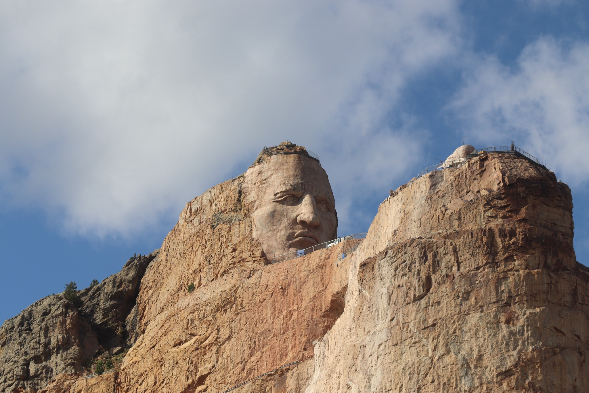 Golden sunrise over pine-covered Black Hills with morning mist weaving through valleys and a colossal sandstone carving of a Native American figure emerging from the cliff face, framed by scaffolding under a bright blue sky.